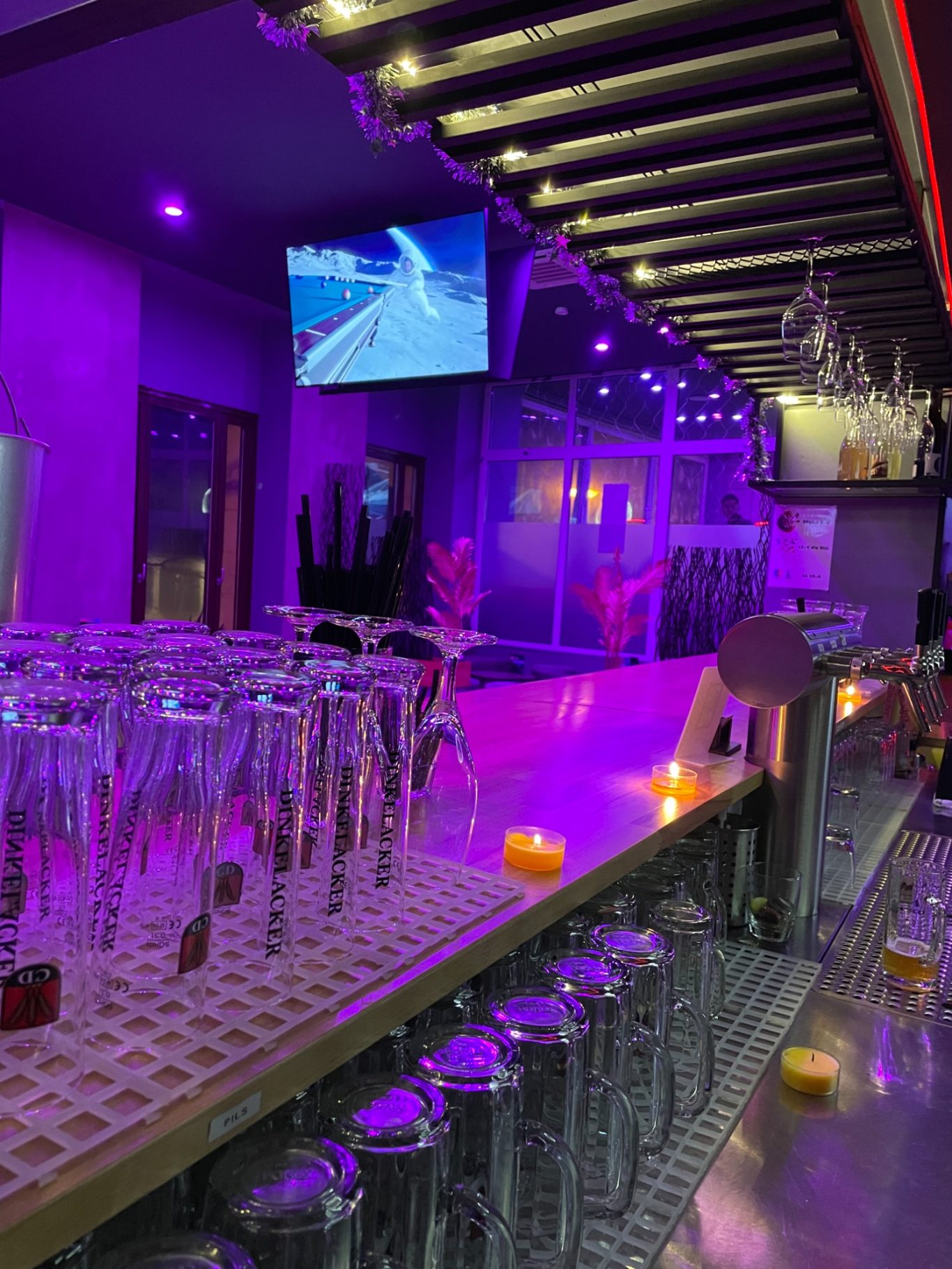Moderne Bar mit lila Beleuchtung, Gläsern und Fernseher im Hintergrund.